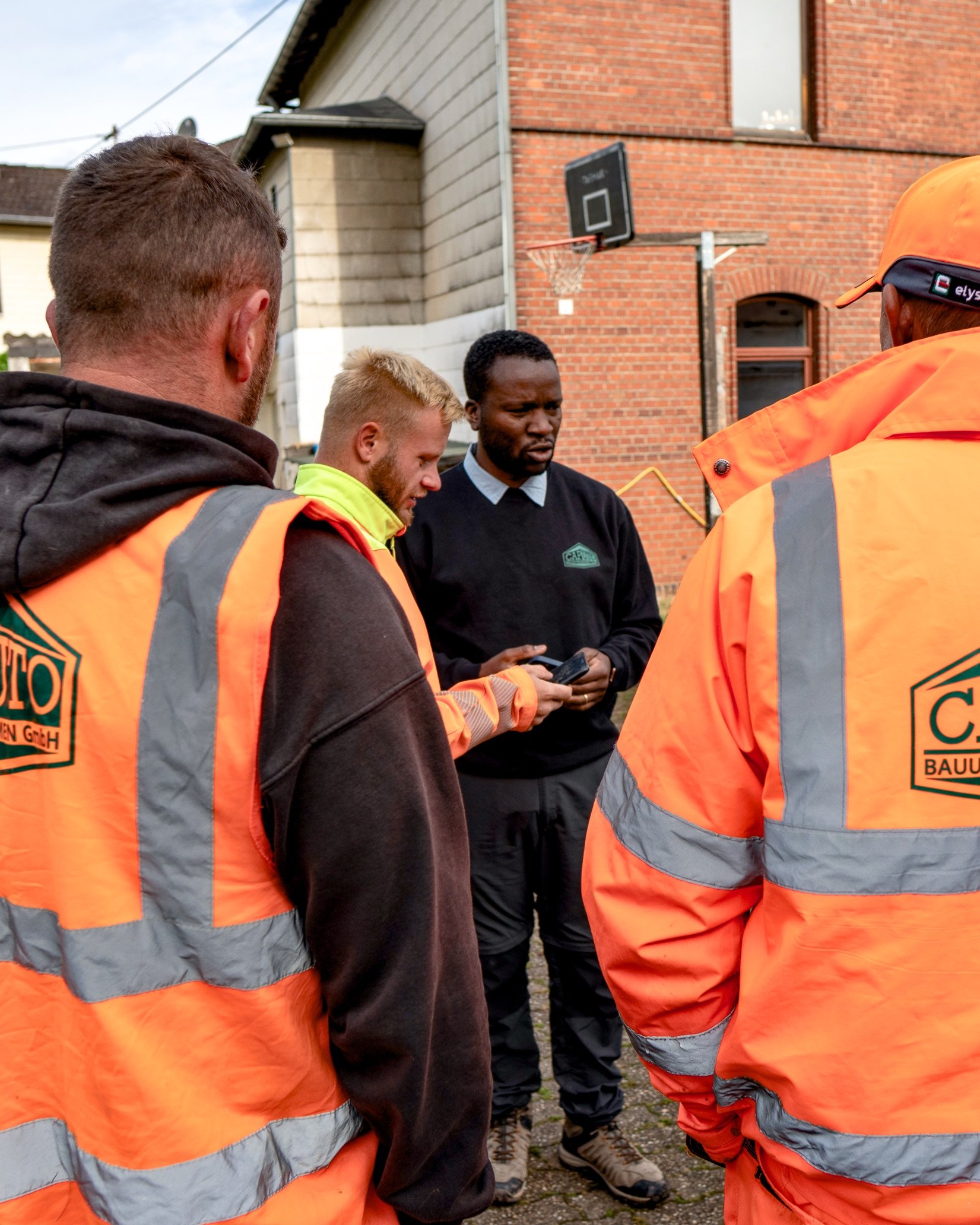 Four construction workers in orange high-visibility jackets discuss a project outdoors, two facing away, while one shows a device to another.