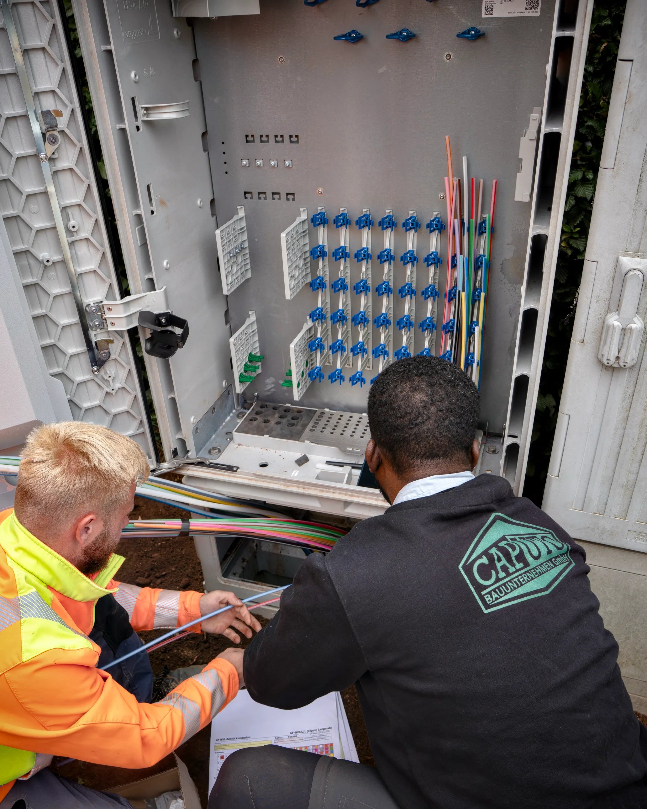 Two technicians work on an open telecom equipment cabinet, examining fiber cables and connectors.