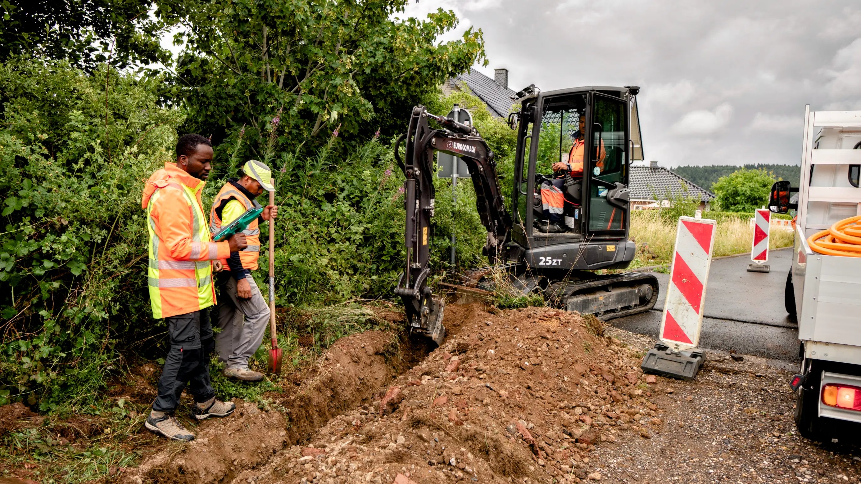 Construction workers operate an excavator and inspect a trench on a roadside with safety barriers and equipment nearby.